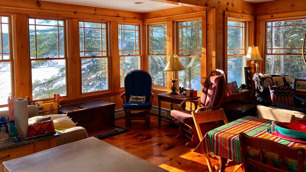A cozy cabin interior with wooden walls and large windows, a dining table, a blue chair, and a colorful tablecloth.