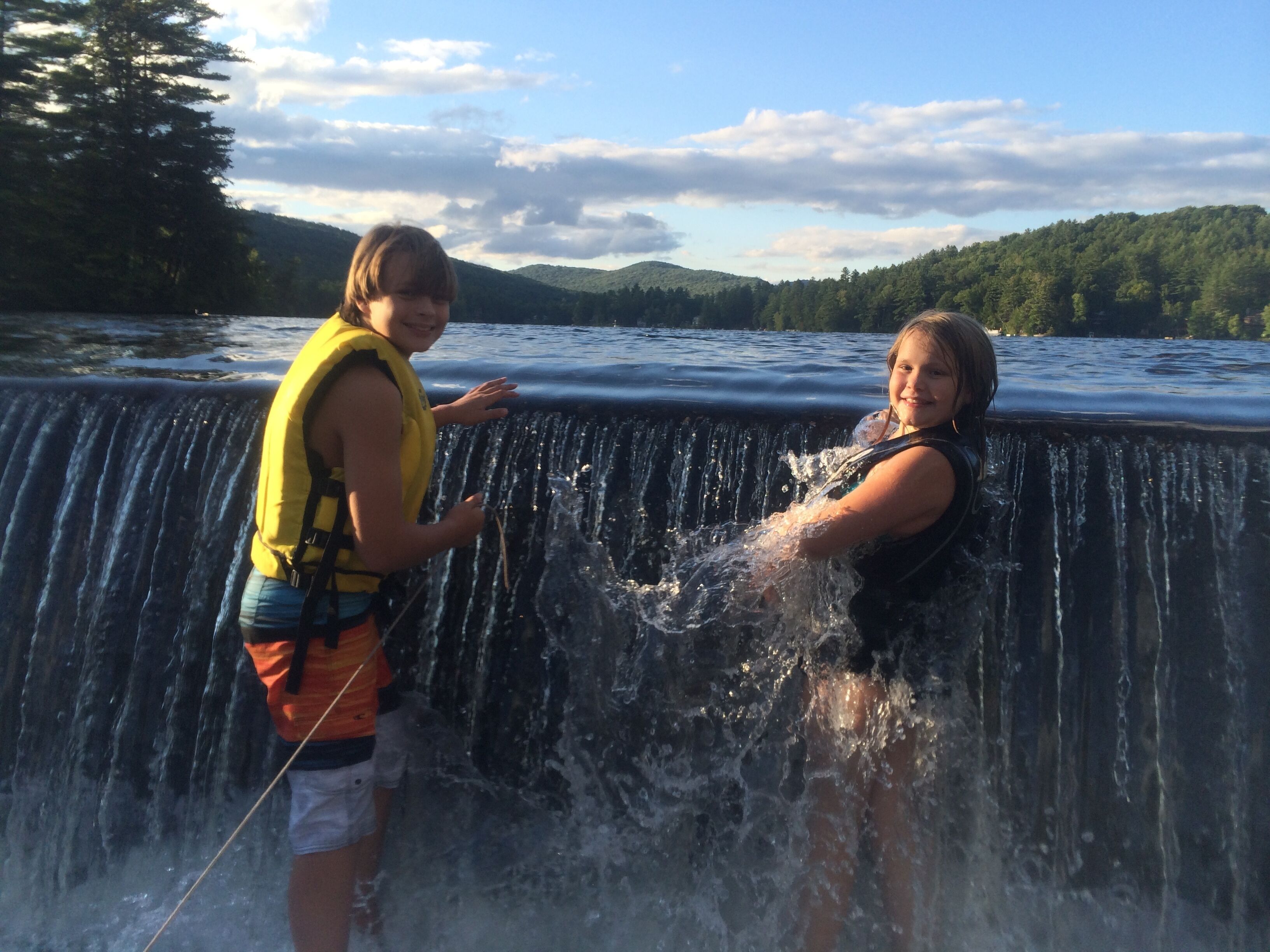 Two people standing at the edge of a water slide, with a lake and forested hills in the background.