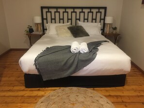 A neatly made bed with white linens, a grey throw blanket, and two rolled towels. There are two bedside tables with lamps and small potted plants. The room has wooden flooring and a round, woven ottoman.