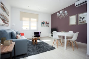 A modern living room with a sofa, a dining table with chairs, a small wooden stool, and a black rug.
