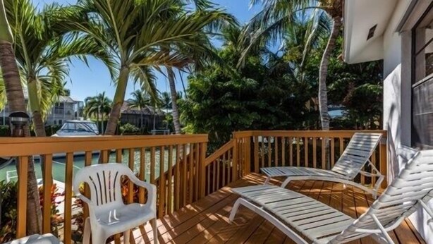 Eine Holzterrasse mit Liegestühlen und einem Plastikstuhl, umgeben von Palmen und einem Pool.