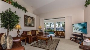 Ein Wohnzimmer mit braunem Ledersofa, einem Couchtisch und einem Fernseher. Ein großes Fenster bietet Blick auf einen Pool und Grünflächen.