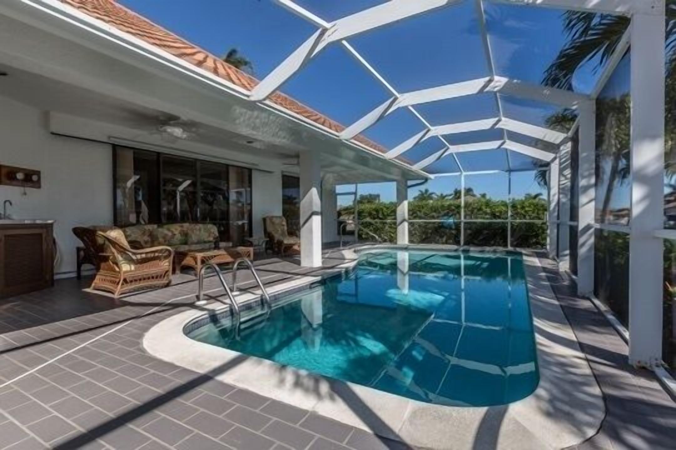 A swimming pool with a glass enclosure, surrounded by outdoor furniture and a covered patio area.
