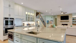 A modern kitchen with a large island, stainless steel appliances, and a staircase leading to a living area with a stone fireplace.