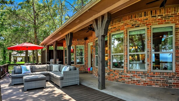 A covered outdoor area with a brick building, large windows, and a wooden deck with outdoor furniture.