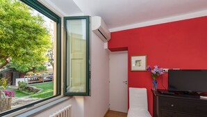 Ein Zimmer mit roter Wand, einem weißen Bett, einem Schminktisch, einem Fernseher und einem Fenster mit Blick auf einen Garten.