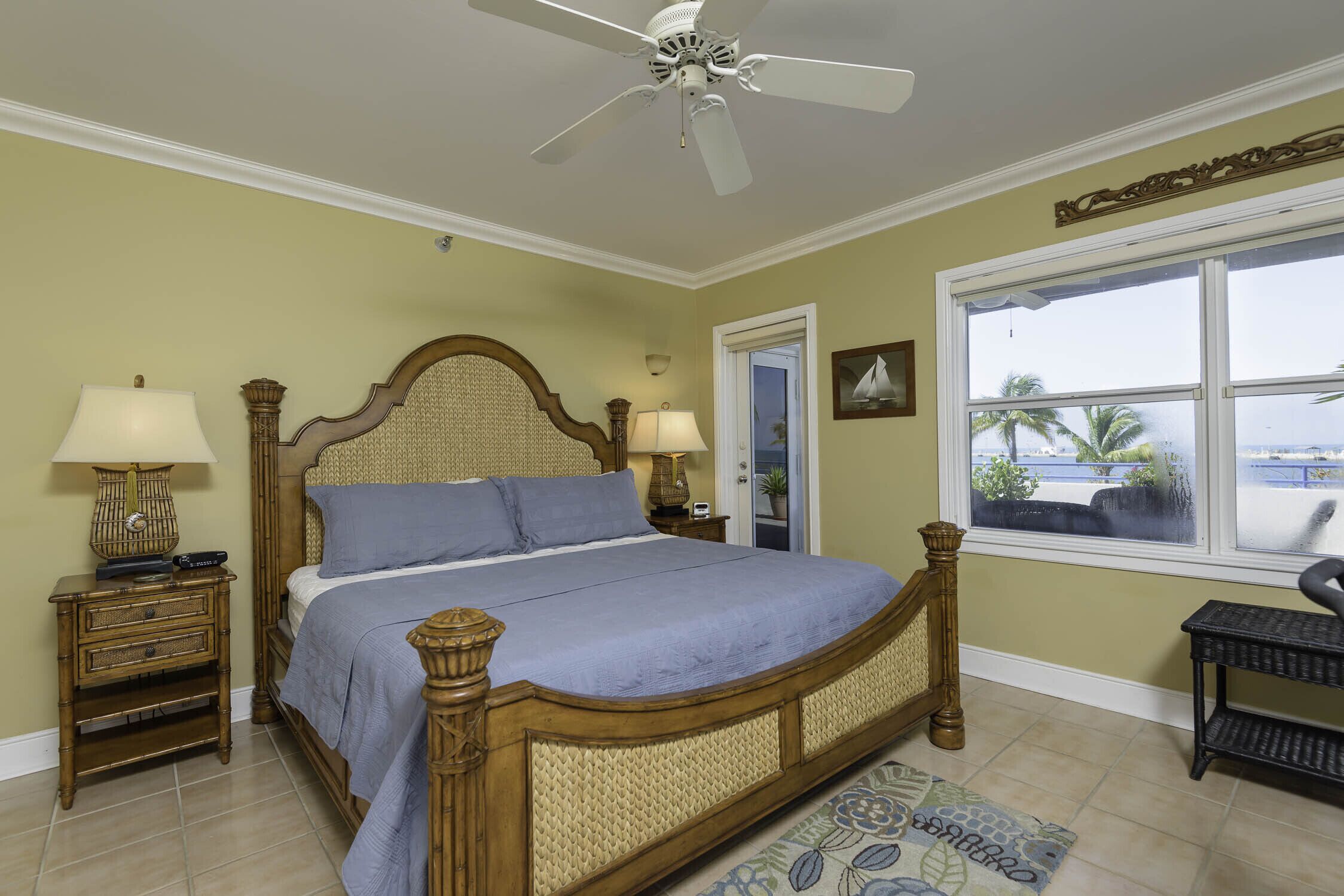A bedroom with a bed, two bedside tables, a ceiling fan, a window with a view, and a door.
