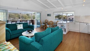 A spacious living area with a sofa, a dining table, and chairs. There is a kitchen area with cabinets and a countertop. Large windows provide natural light and views of the outdoors.