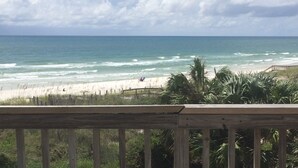 Une vue sur la plage avec une terrasse en bois au premier plan, des vagues de l’océan et un ciel dégagé.