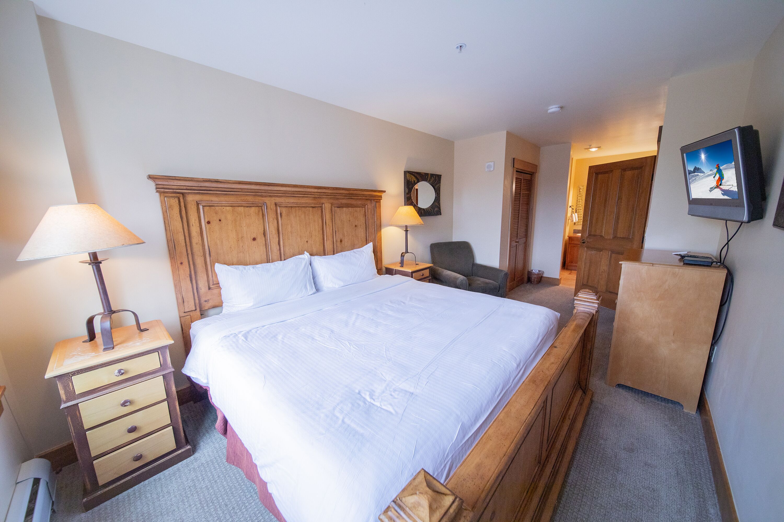 A hotel room with a large bed, wooden headboard, a desk with a lamp, a chair, a TV, and a wardrobe.
