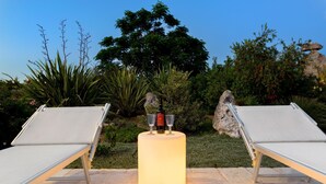 Deux chaises longues, séparées par une structure lumineuse cylindrique, sont disposées sur une terrasse en pierre, entourée de verdure et de rochers.