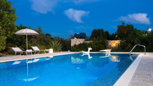 Une piscine aux eaux cristallines, bordée de chaises longues et agrémentée d’un parasol, le tout entouré de verdure et d’un mur de pierre.