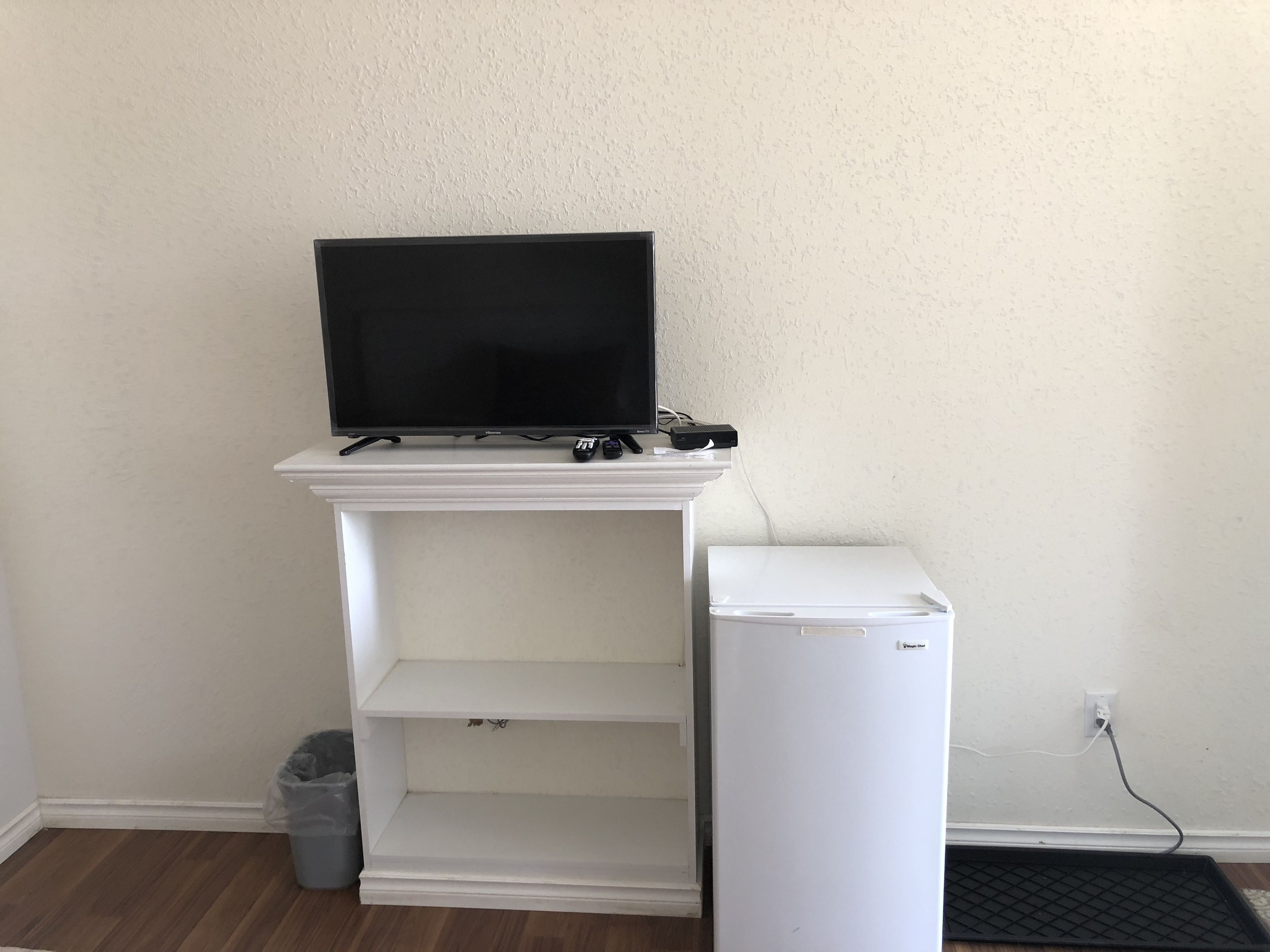 Un mueble blanco de entretenimiento con un televisor de pantalla plana, un refrigerador blanco pequeño y un cubo de basura.