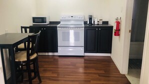 A compact kitchen with a microwave, stove, and coffee maker. There is a dining table with chairs and a fire extinguisher mounted on the wall. - Langham Motel (Langham)