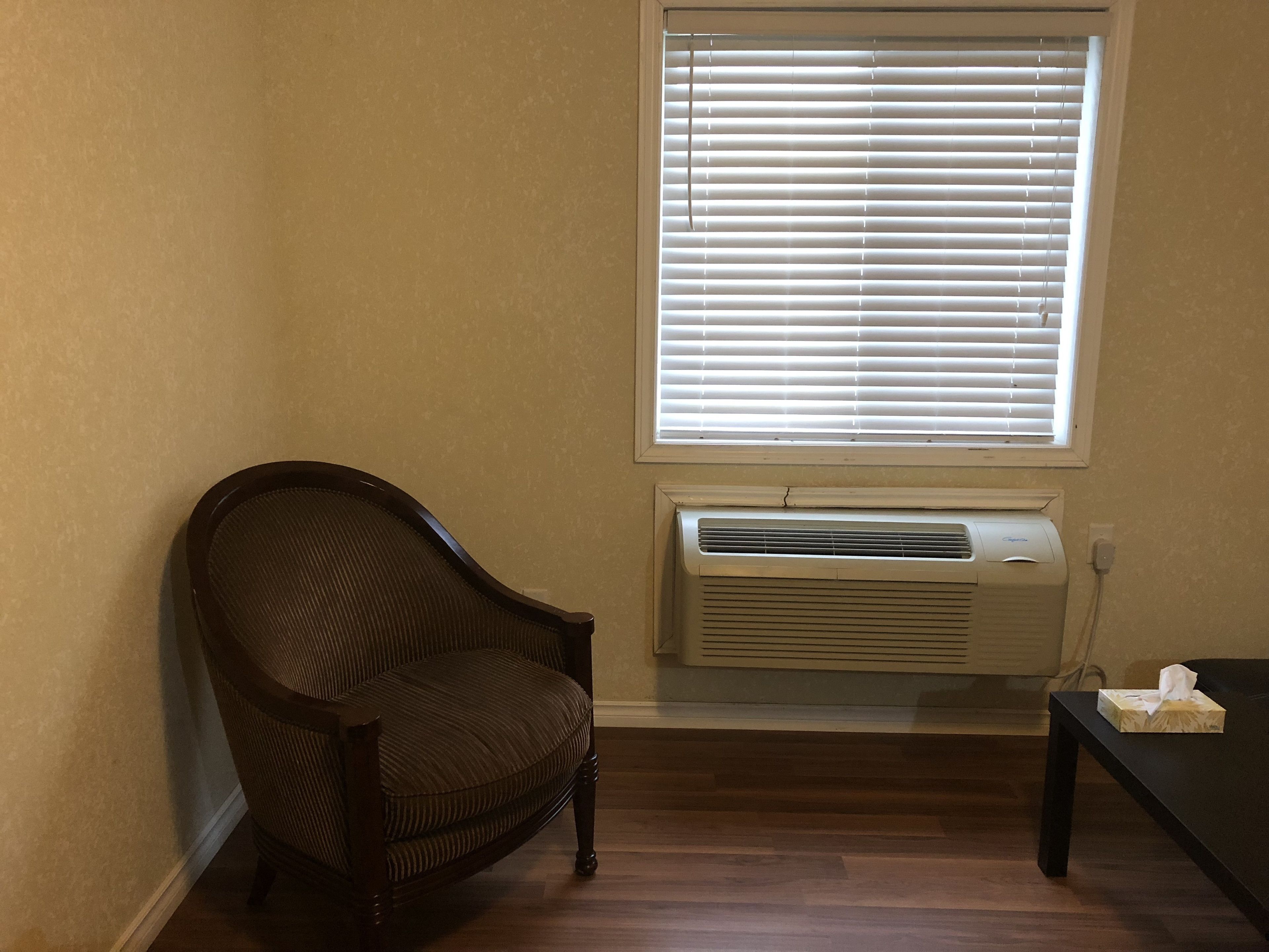 A corner room with a dark brown chair, a window with blinds, an air conditioning unit, and a small table with a tissue box.