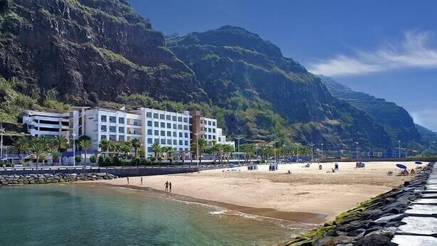 Et kysthotel med udsigt til stranden, en klippefyldt kystlinje og et bjerglandskab i baggrunden.