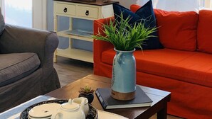 A living room with a red sofa, a grey armchair, a small table with a teapot and plates, a blue vase with green plants, and a white cabinet with decorative items. - Sunny Days - Beautiful, large Deck, and Nice Views! Free WIFI & Pet-Friendly! (Surfside Beach)