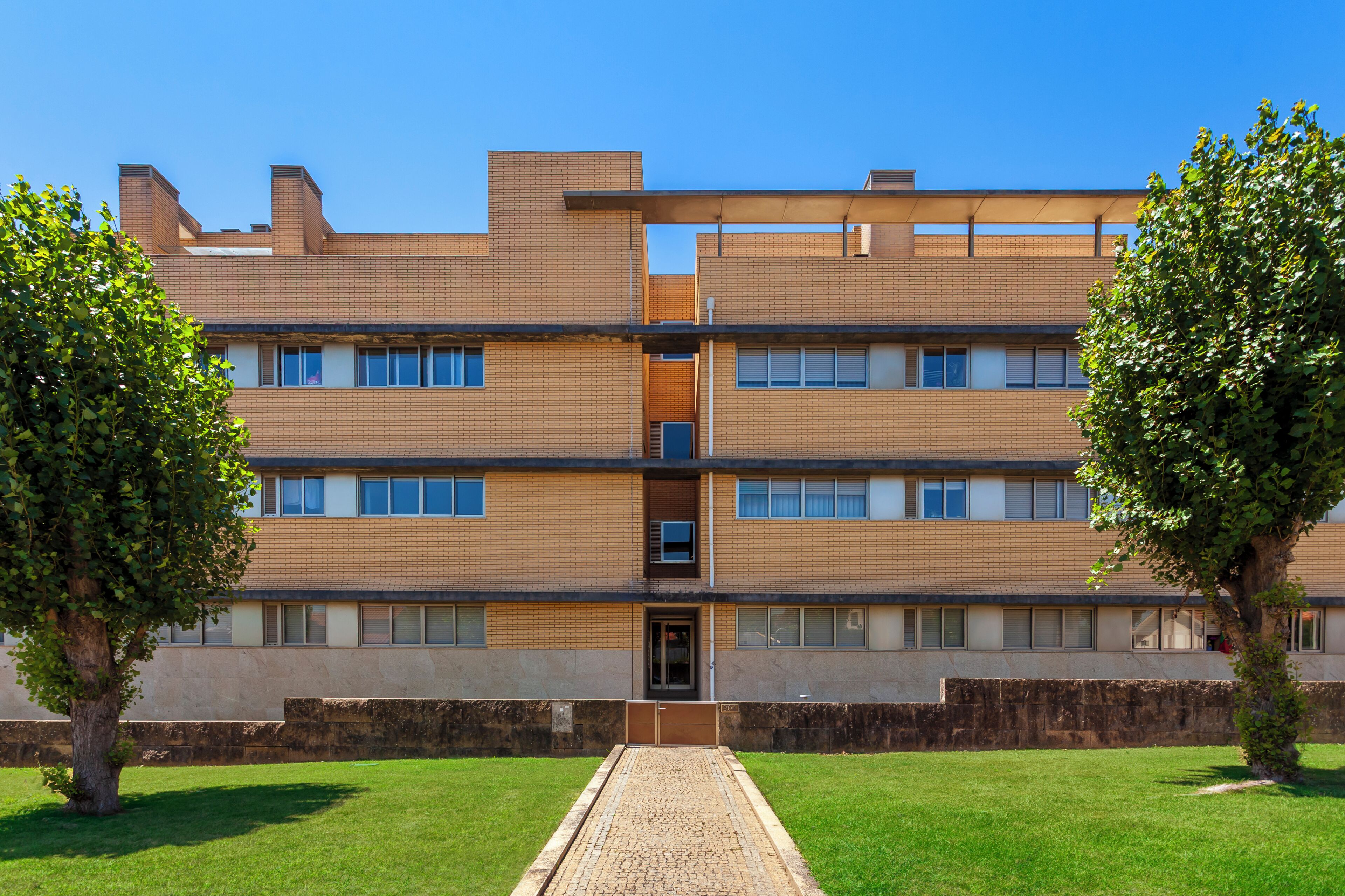 Um edifício de vários andares com fachada simétrica, entrada central e um caminho pavimentado que leva à entrada.