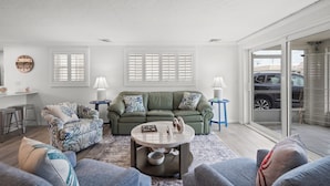 A spacious living room with a green sofa, a patterned armchair, and a round coffee table. There are two side tables with lamps, a large window with shutters, and a view of parked cars outside. - Island House Beach Resort V5 (Siesta Key)