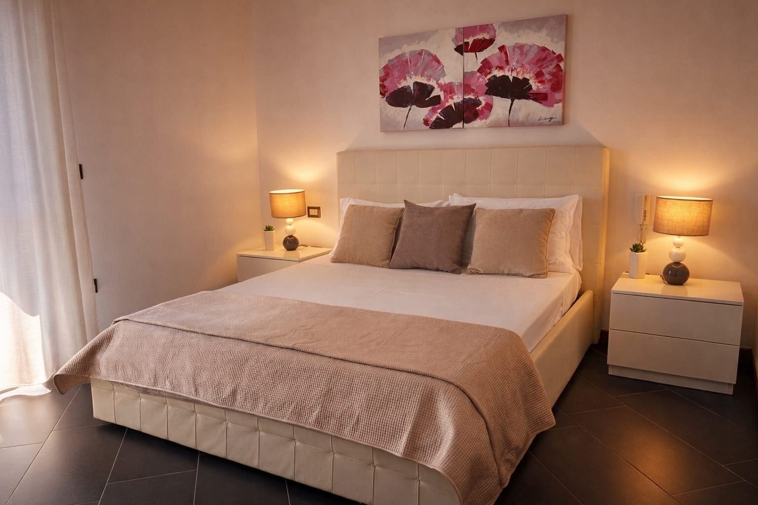 Un dormitorio con una cama grande, dos mesitas de noche con lámparas, una obra de arte en la pared y una ventana con cortinas.