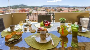 Un tavolo da balcone apparecchiato con articoli per la colazione, tra cui frutta, una brocca e bicchieri, con vista su una città con edifici a tetti rossi e colline sullo sfondo.