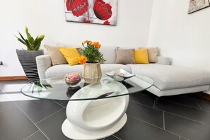 A modern living room with a glass coffee table, a sofa with cushions, a potted plant, and a wall-mounted artwork.