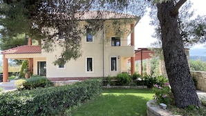 Un edificio de dos pisos con jardín, un árbol grande y un muro de piedra.