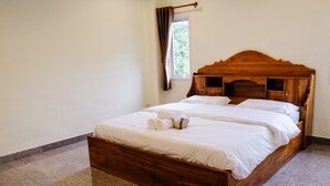 Un lit en bois avec du linge de lit blanc, une fenêtre avec des rideaux et une petite table de chevet sur laquelle se trouvent une serviette et un panier.