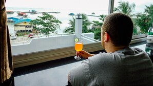 A person sitting by a window with a view of a coastal area, holding a drink with a lime garnish. - The Lake House Ko Yo (Songkhla)