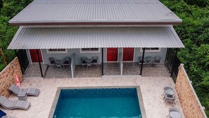 Une piscine rectangulaire dont les bords sont recouverts de carreaux, entourée de chaises longues et d’un espace de repos abrité avec des portes rouges.