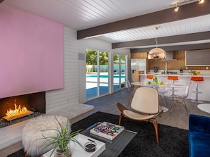 A modern living room with a fireplace, a pool view, and a kitchen area.