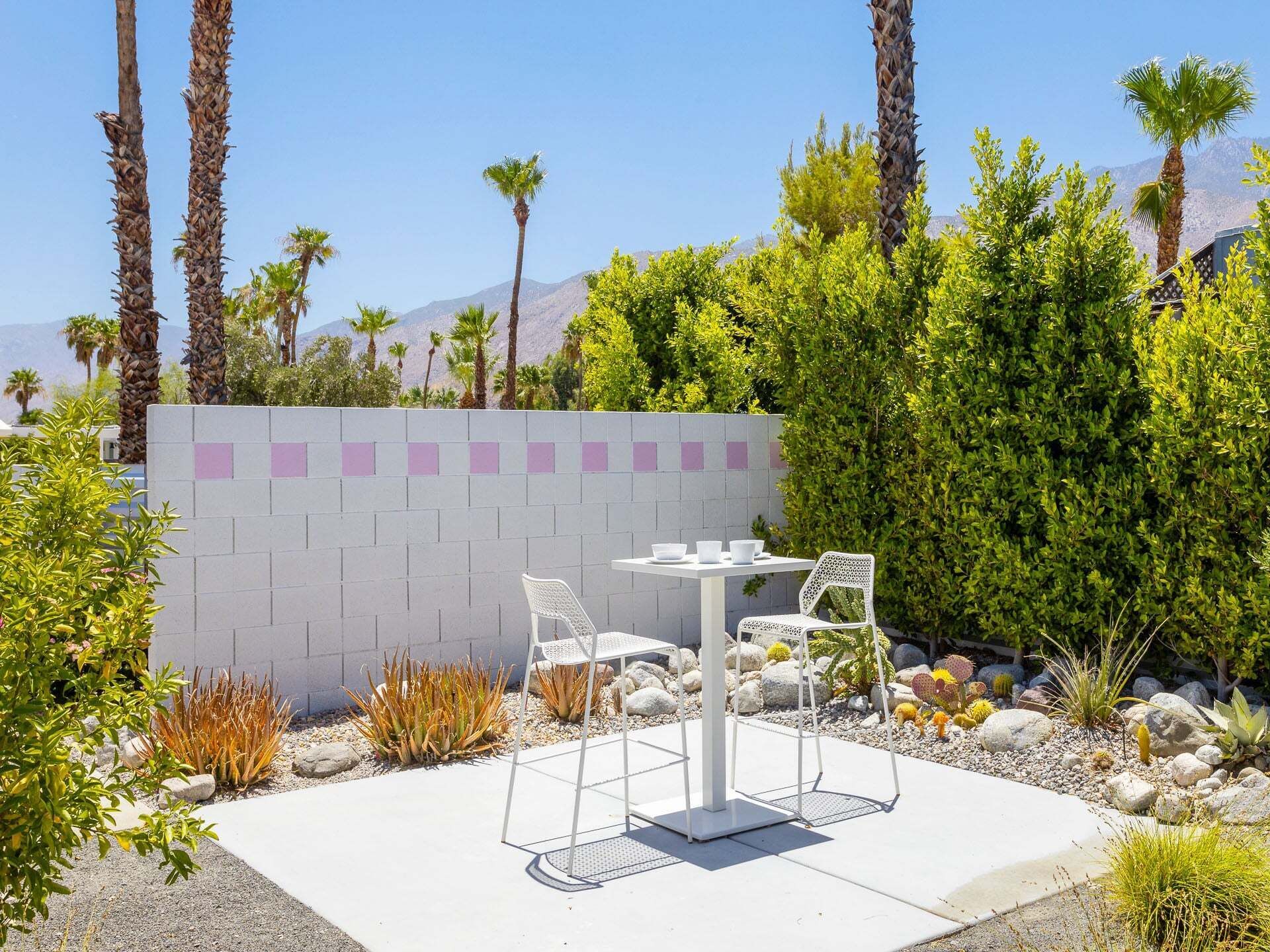 Een terras met een witte tafel en stoelen, omringd door diverse planten en een witte muur met roze accenten.