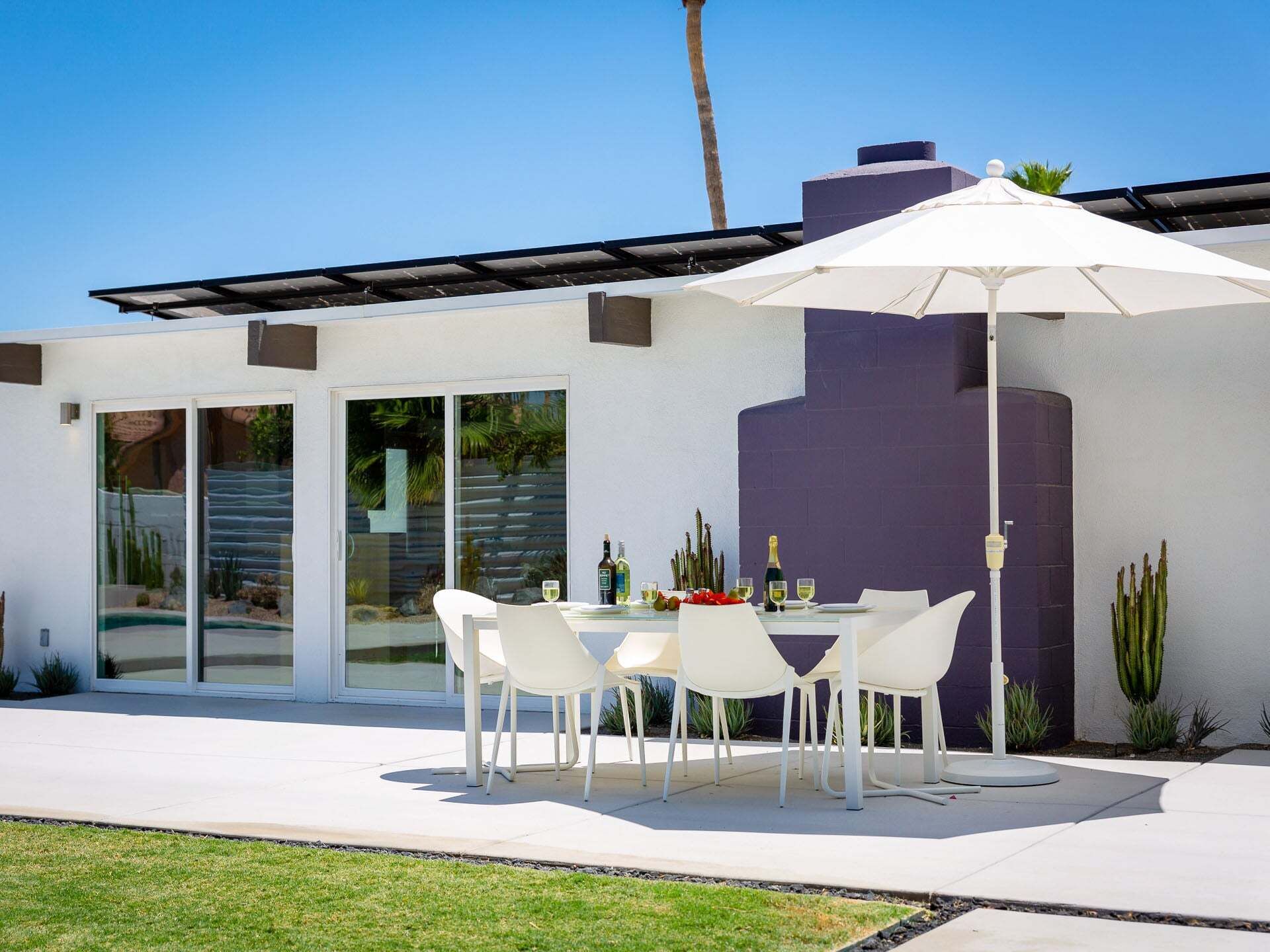 Une terrasse avec une table et des chaises, un parasol blanc et un bâtiment moderne doté de portes vitrées et d’un mur violet.