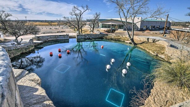 A clear blue swimming pool with stone walls and a few red and white buoys, surrounded by a stone pathway and sparse trees.