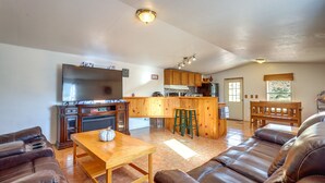 A living room with a sofa, a wooden table, and a TV. There is a kitchen area with wooden cabinets and a bench.