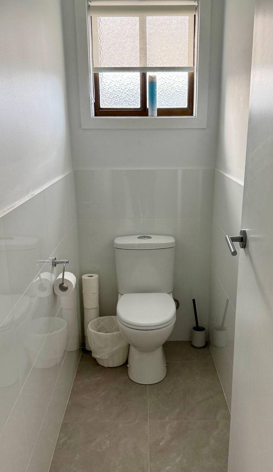 Une petite salle de bain propre, équipée de toilettes, d’un rouleau de papier toilette, d’une poubelle et d’une fenêtre située au-dessus des toilettes.