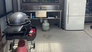 A kitchen with a gas stove, a gas cylinder, a refrigerator, and a window with a view of trees. - Beach House Getaway #2 (Smiths Beach)