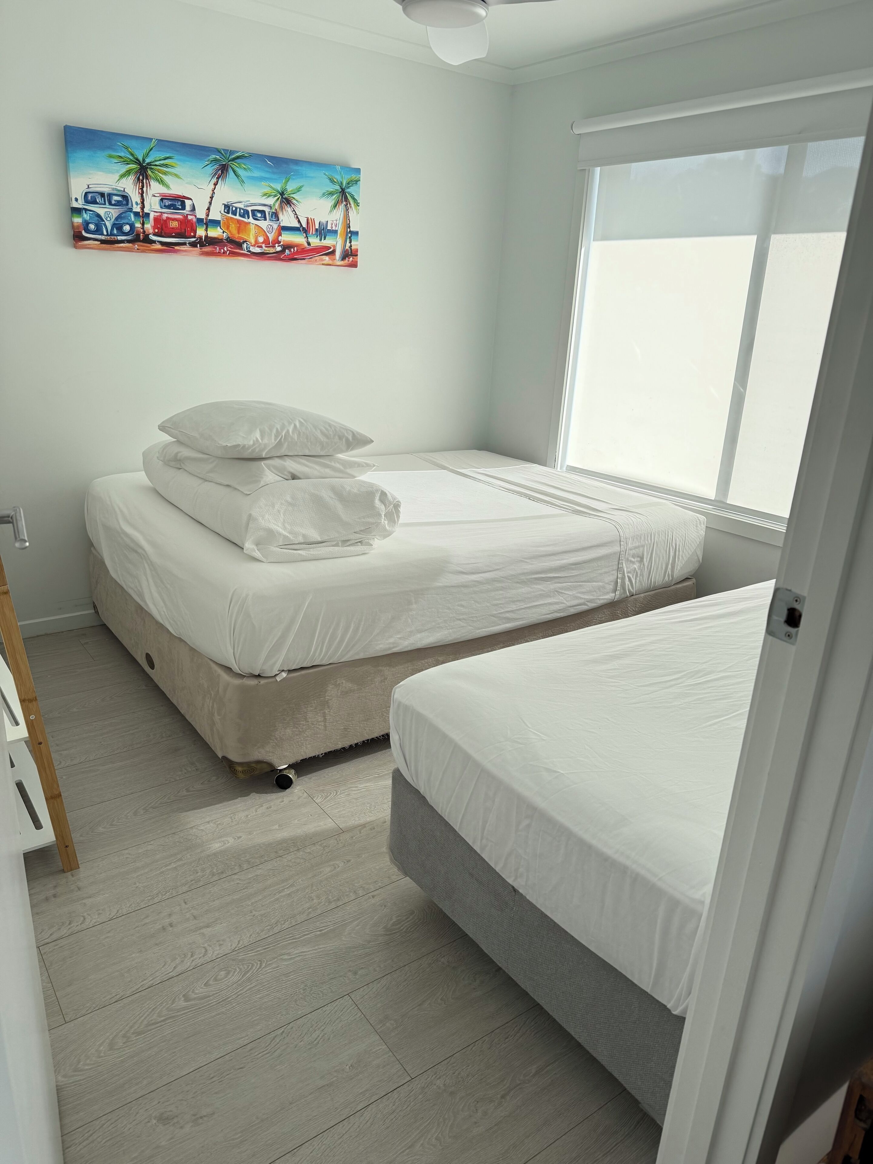 Une chambre avec deux lits, un tableau représentant des fourgonnettes sur une plage et une grande fenêtre.