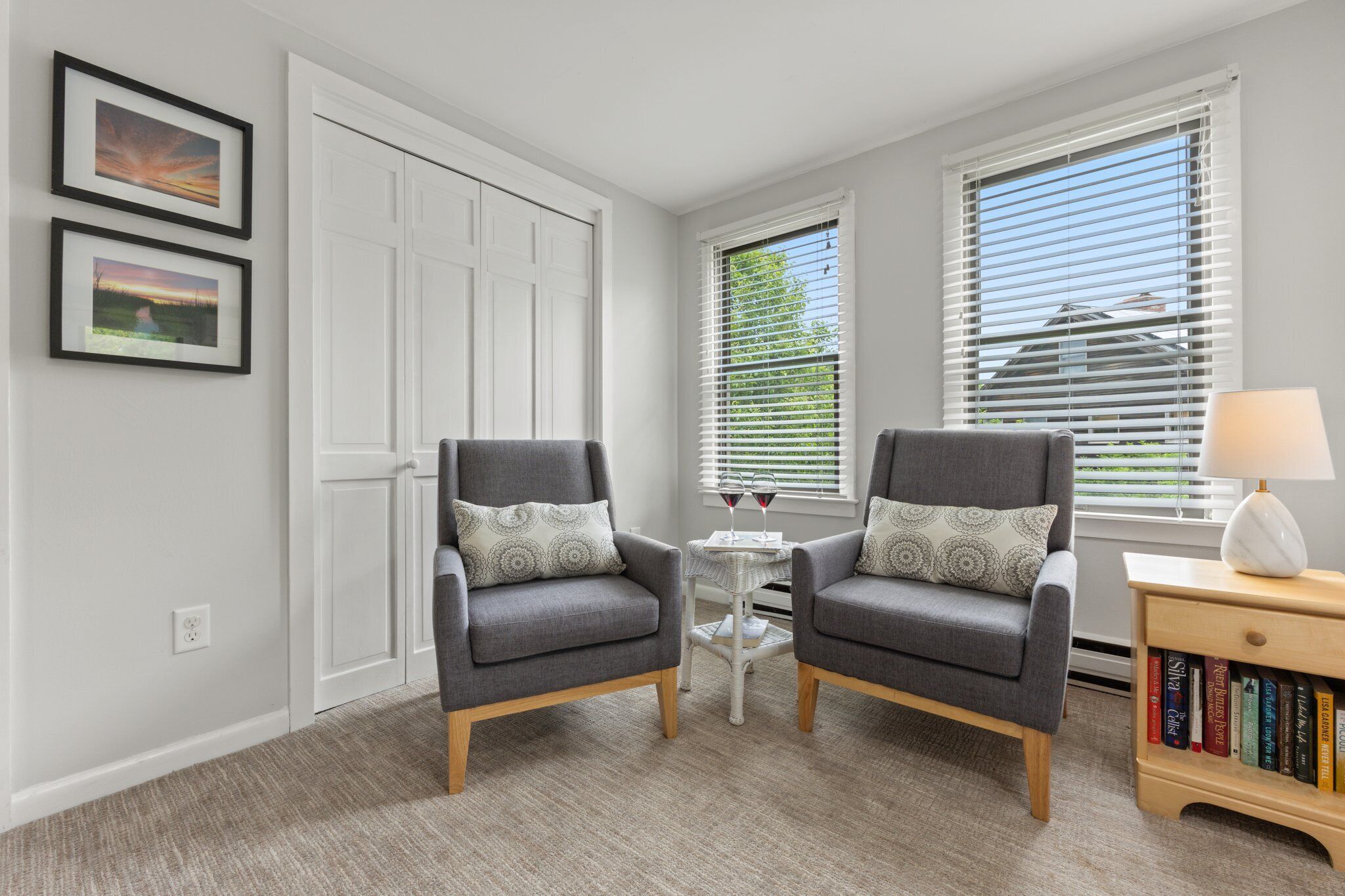 A room with two grey armchairs, a small round table with a lamp, and a bookshelf.