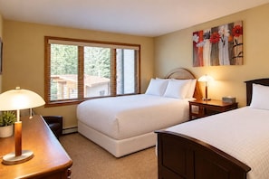 A hotel room with two beds, a desk with a lamp, a painting on the wall, and a window with blinds.