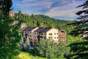 A multi-story residential building with a sloped roof, surrounded by greenery and a mountainous backdrop.