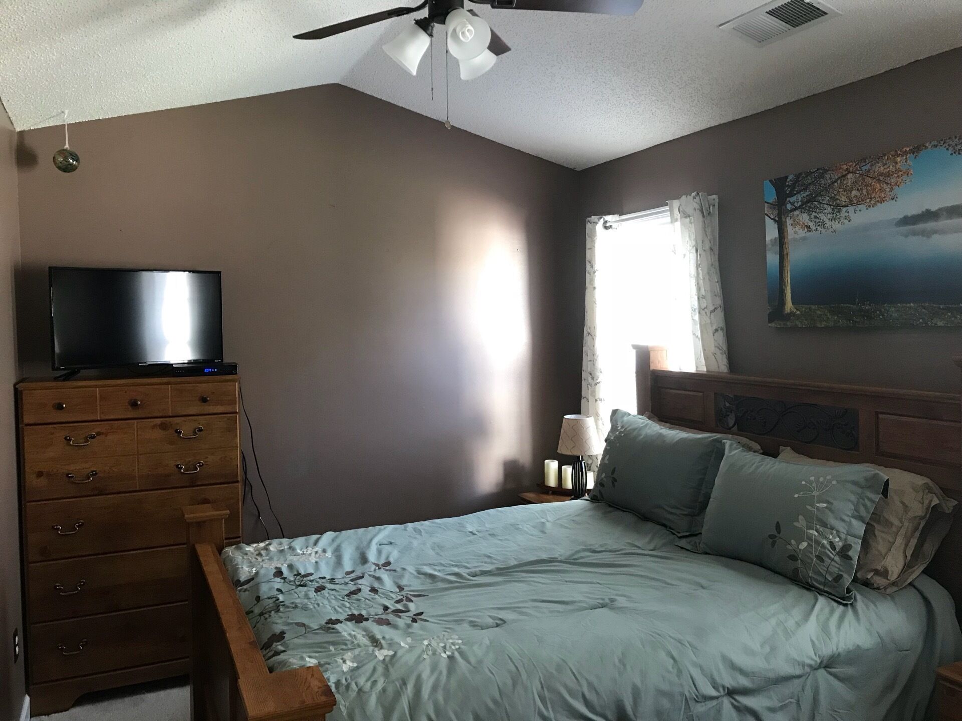 Une chambre à coucher avec un lit, une commode, un ventilateur de plafond et un tableau accroché au mur.