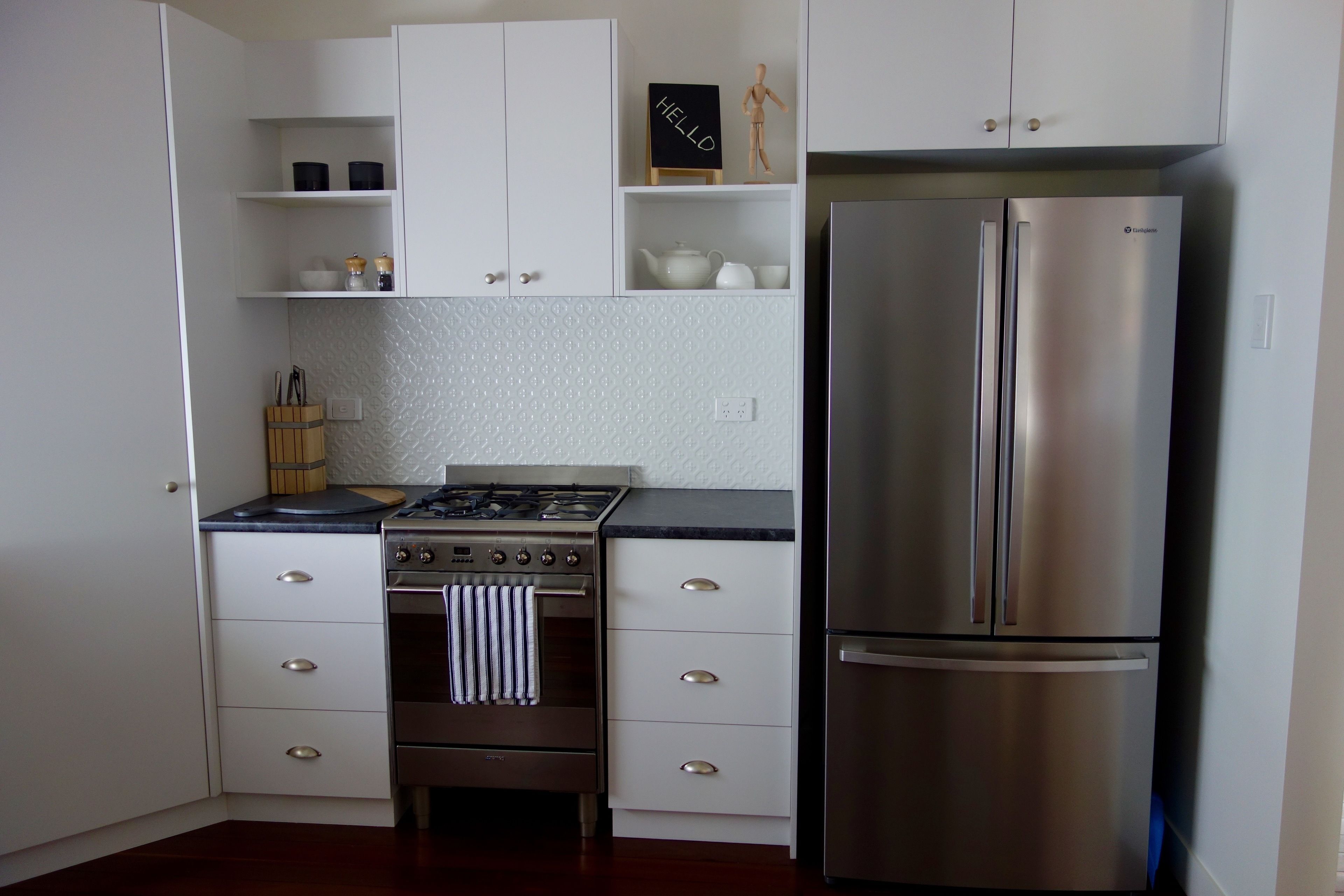 A kitchen with white cabinets, a stainless steel refrigerator, a gas stove, and a small wooden shelf with a 'HELLO' sign.