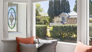 Ein Zimmer mit großem Fenster, einer Bank mit Kissen und Blick auf Garten und Gebäude.