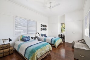 A bedroom with two beds, a ceiling fan, a window with blinds, and a door leading to another room. - The Lighthouse (East Toowoomba)