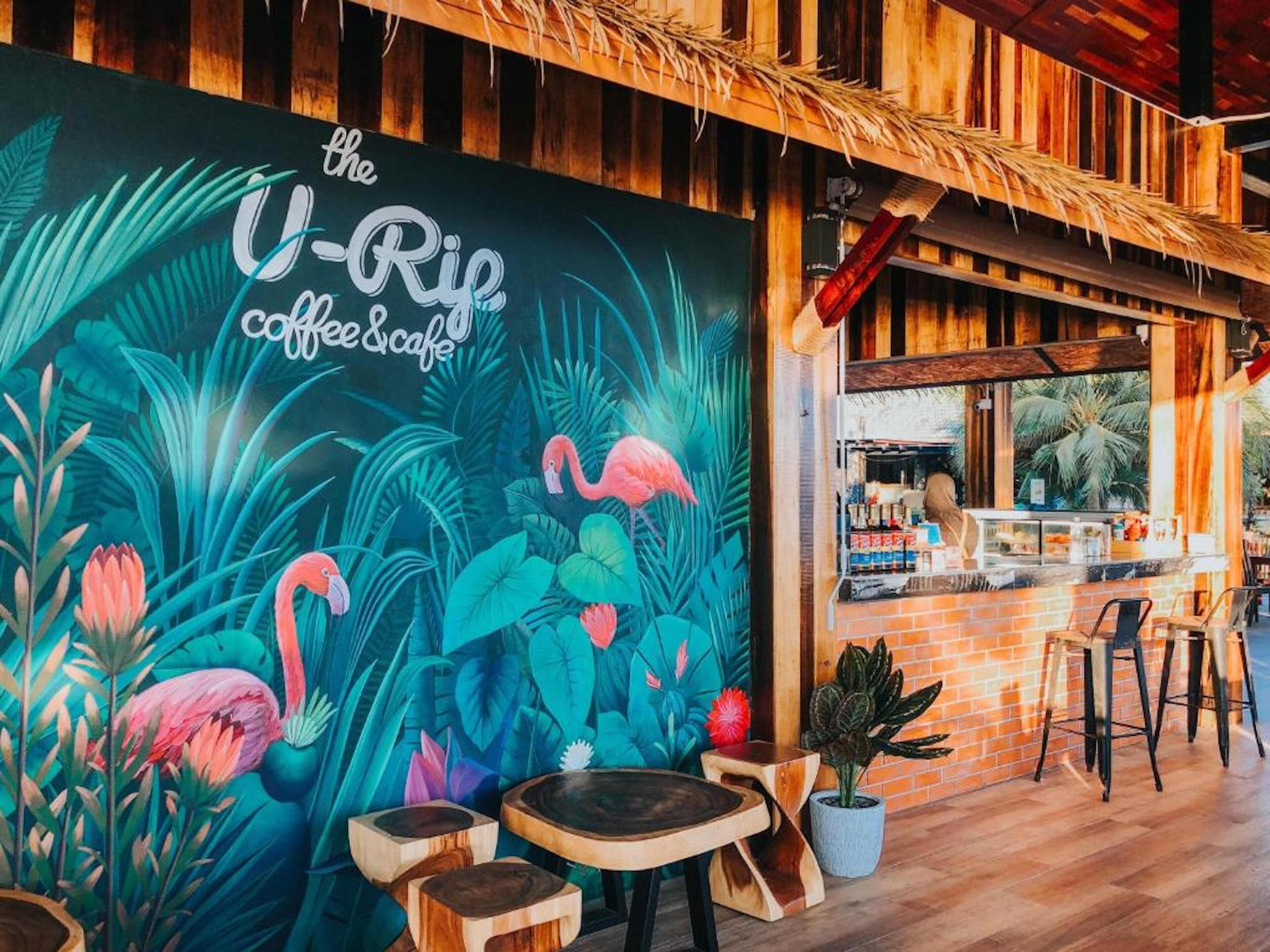 Een levendige muurschildering met tropische flora en flamingo's, een rustieke houten bar en houten tafels en stoelen.