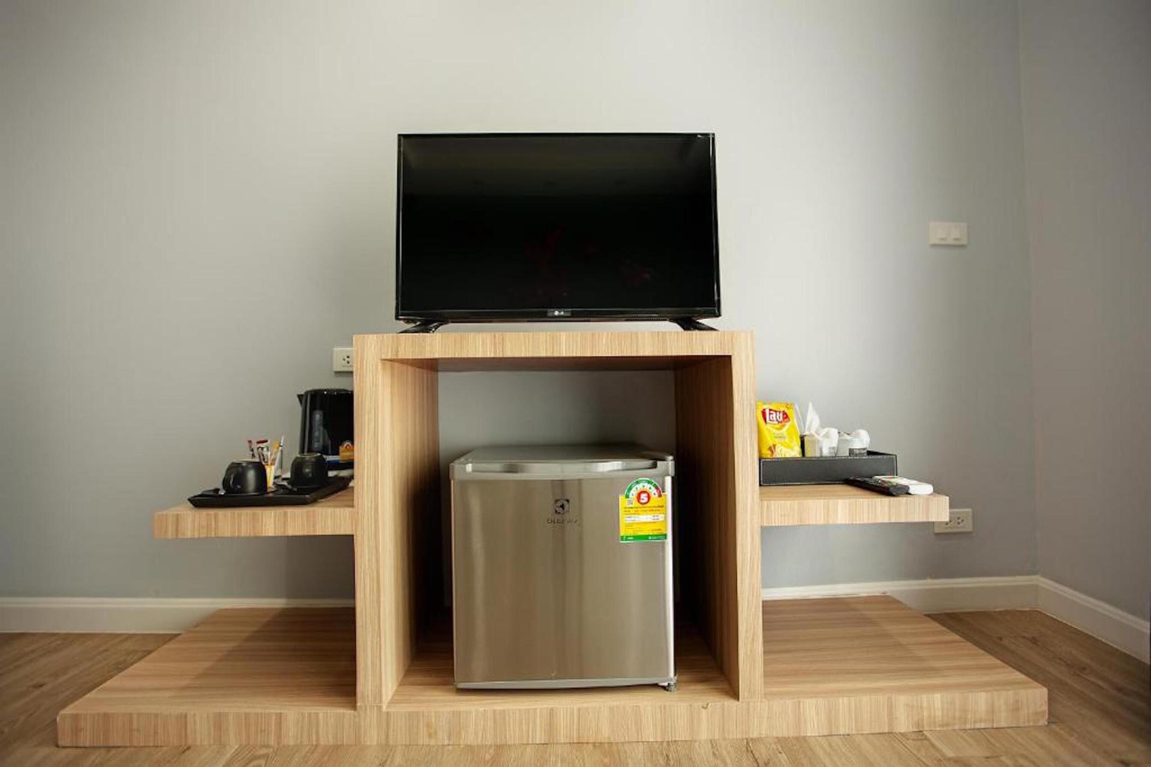A compact living space with a flat-screen TV on a wooden stand, a stainless steel mini-fridge, and a shelf with a coffee maker and snacks.