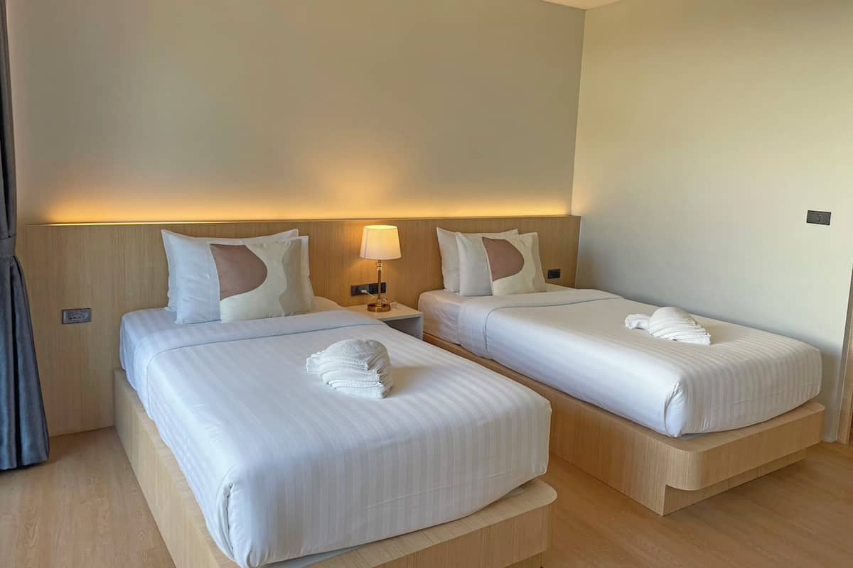 Quarto de hotel com duas camas de solteiro, cada uma com cabeceira de madeira, mesa de cabeceira com abajur e iluminação embutida na parede.