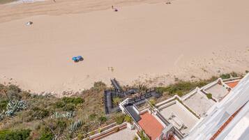 Luchtbeeld van een strand met een heldere lucht, rustige zee en enkele boten. Er staan gebouwen met rode daken en een verharde promenade langs het strand.