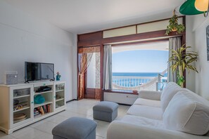 A living room with a sofa, ottoman, TV, and a bookshelf. There is a large window with a view of the sea. - Direct & Private beach access. 30 km from Lisbon (Sesimbra)
